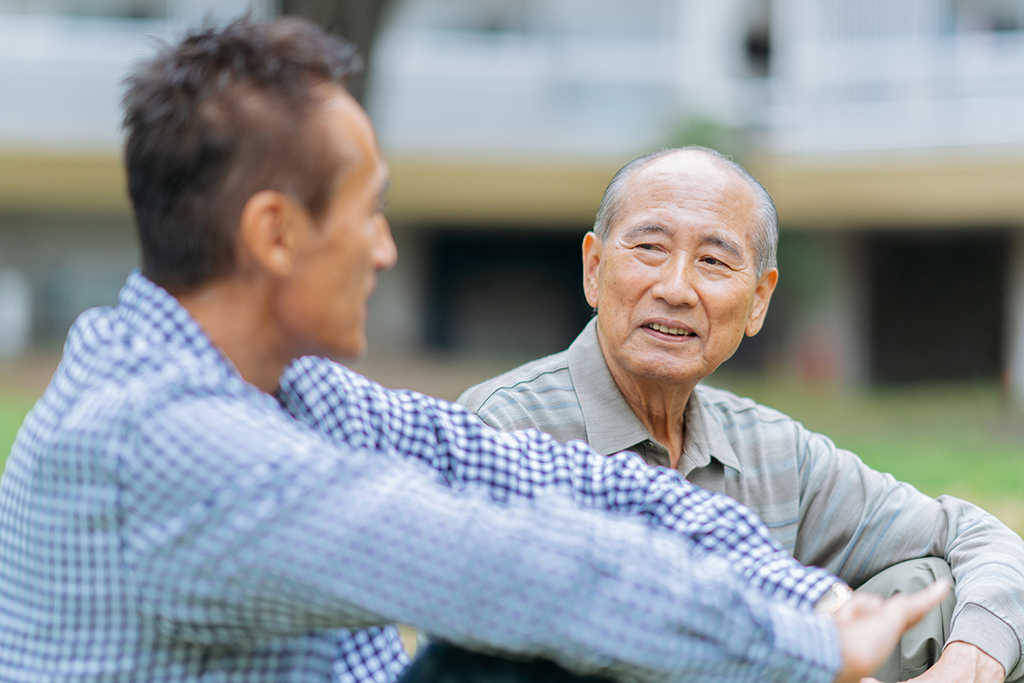 A senior adult man is enjoying talking with his son in a public park outdoors.