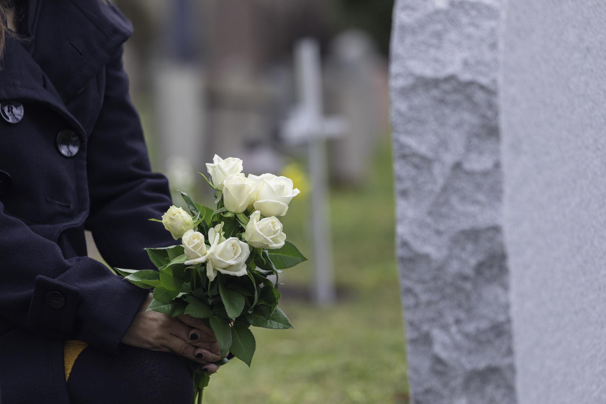 A woman kneeling by a grave at the cemetery bringing a bouquet of flowers.