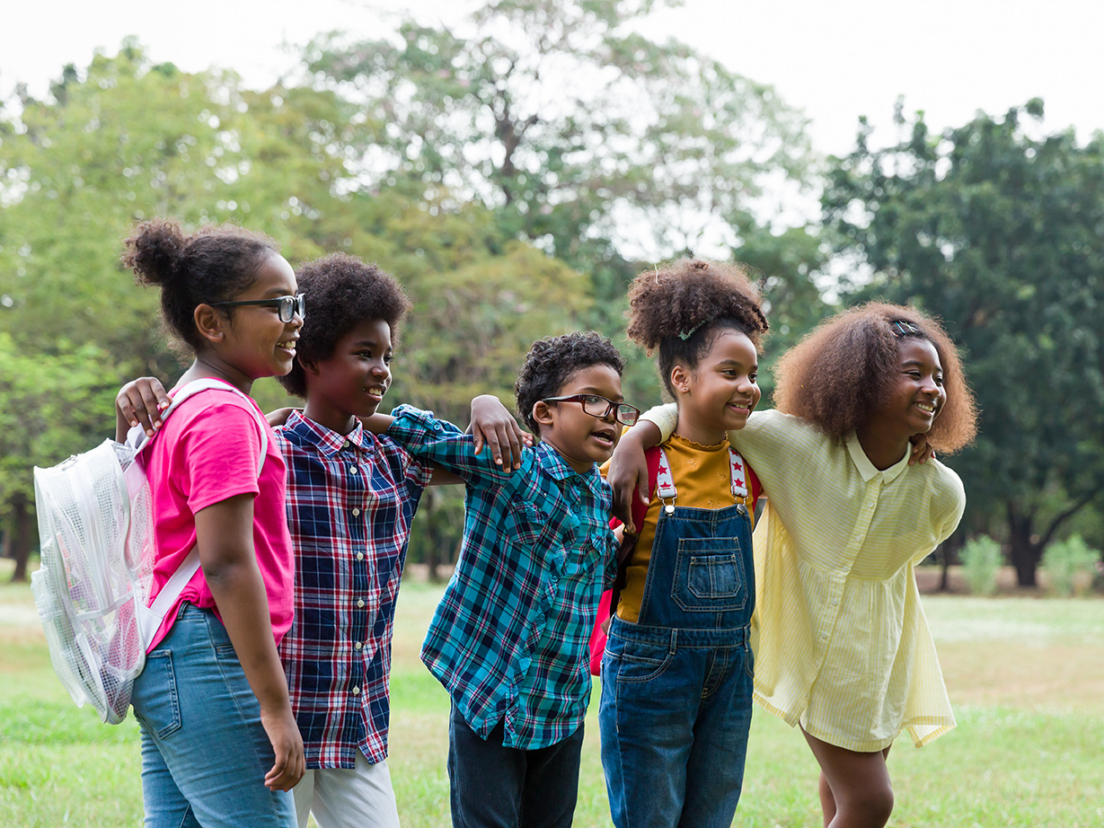 Five children smiling and standing together in a park, with trees in the background.