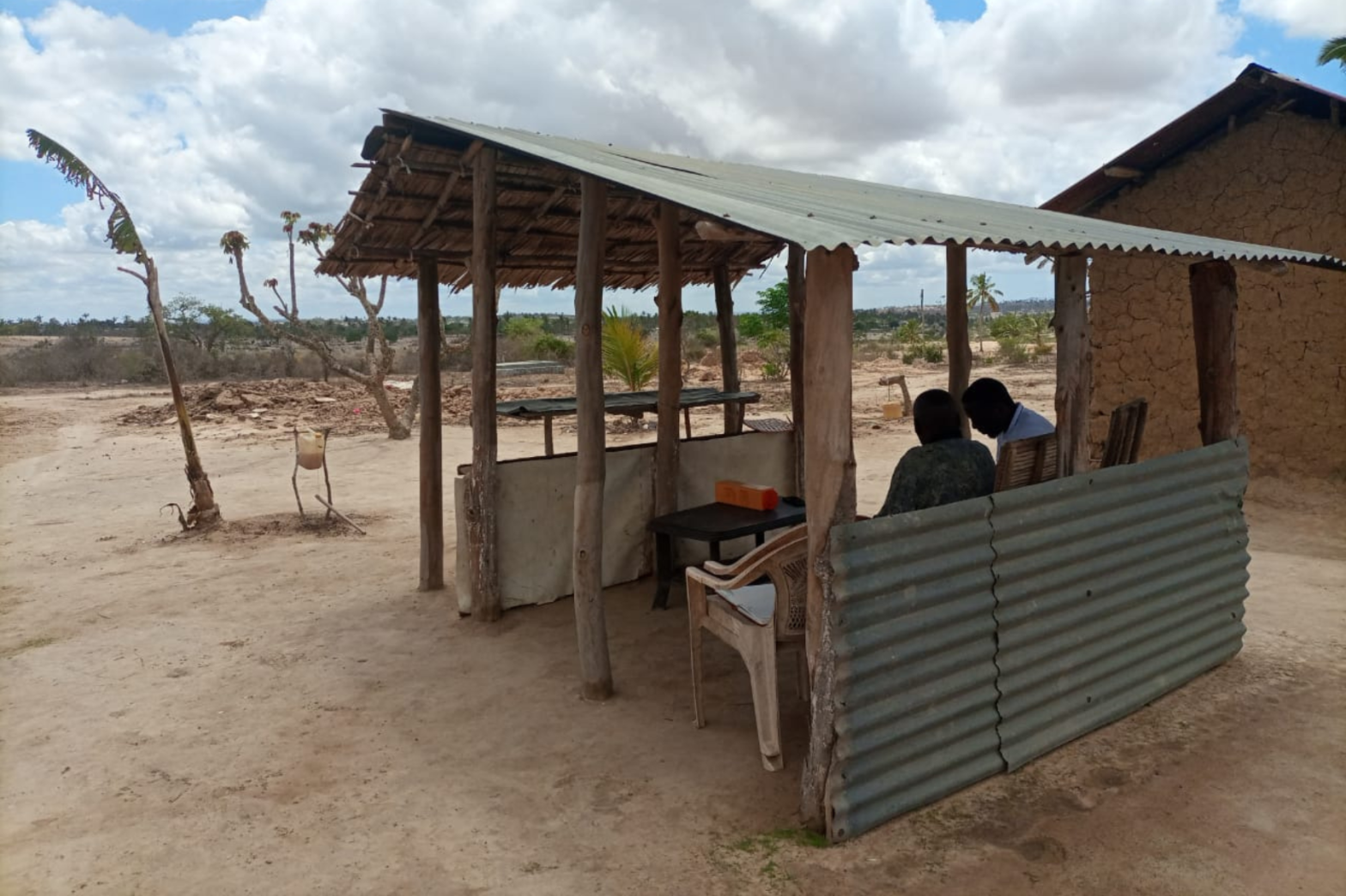 Outdoor shelter with corrugated metal roof, two people seated inside, dusty terrain, and a mud-brick building nearby.