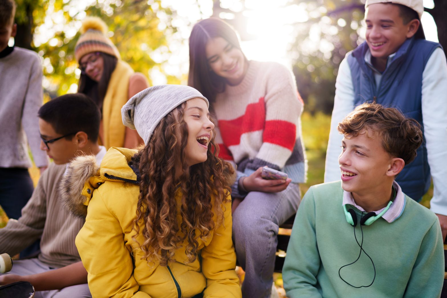 Happy teenage boy and girl laughing while sitting whit their friends on the bench during autumn day