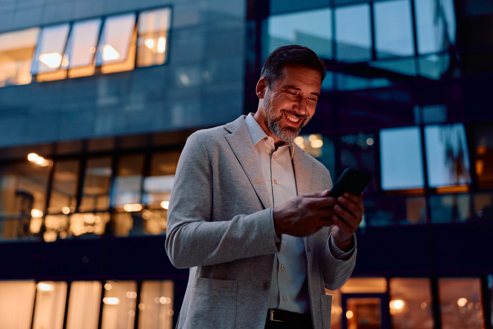 Smiling man standing against a glowing office building, happily engaged with his mobile phone.