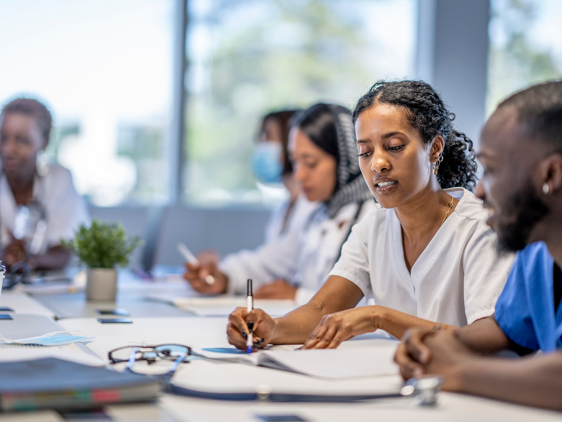 A small group of medical professionals sit around a boardroom table as they meet to discuss patient cases. They are each dressed professionally in scrubs and lab coats as they work through the case files spread out on the desk collaboratively in small groups.