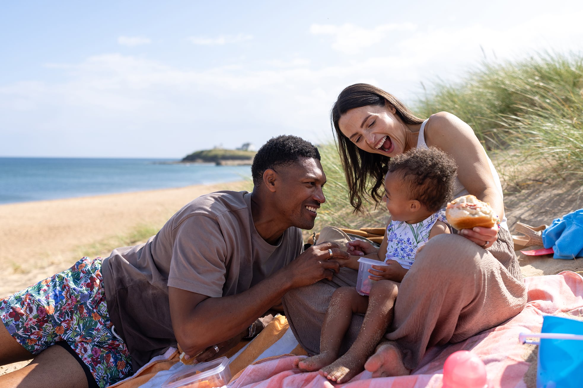 A medium shot of a joyful family enjoying a relaxing beach picnic on a sunny summer day. The young parents sit on a colourful blanket with their toddler, surrounded by beach toys and picnic items. The father holds food for the child while the mother watches with a loving smile.