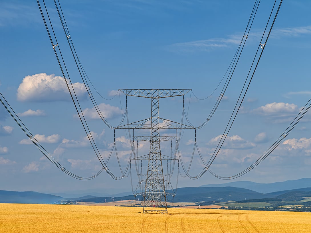 High voltage power lines and transmission towers stretch across a golden wheat field toward distant hills under a bright blue sky.