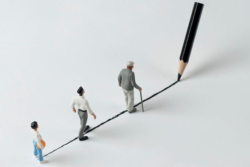 Three miniature figurines walking along a drawn black line, with a large pencil at one end. The figures include a child, a man, and an elderly man with a cane.