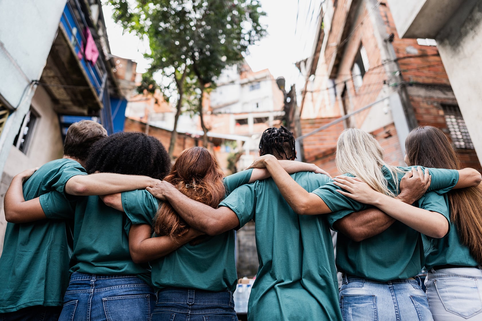 A group of seven people in green T-shirts stands with arms around each other, facing away in an urban neighborhood.