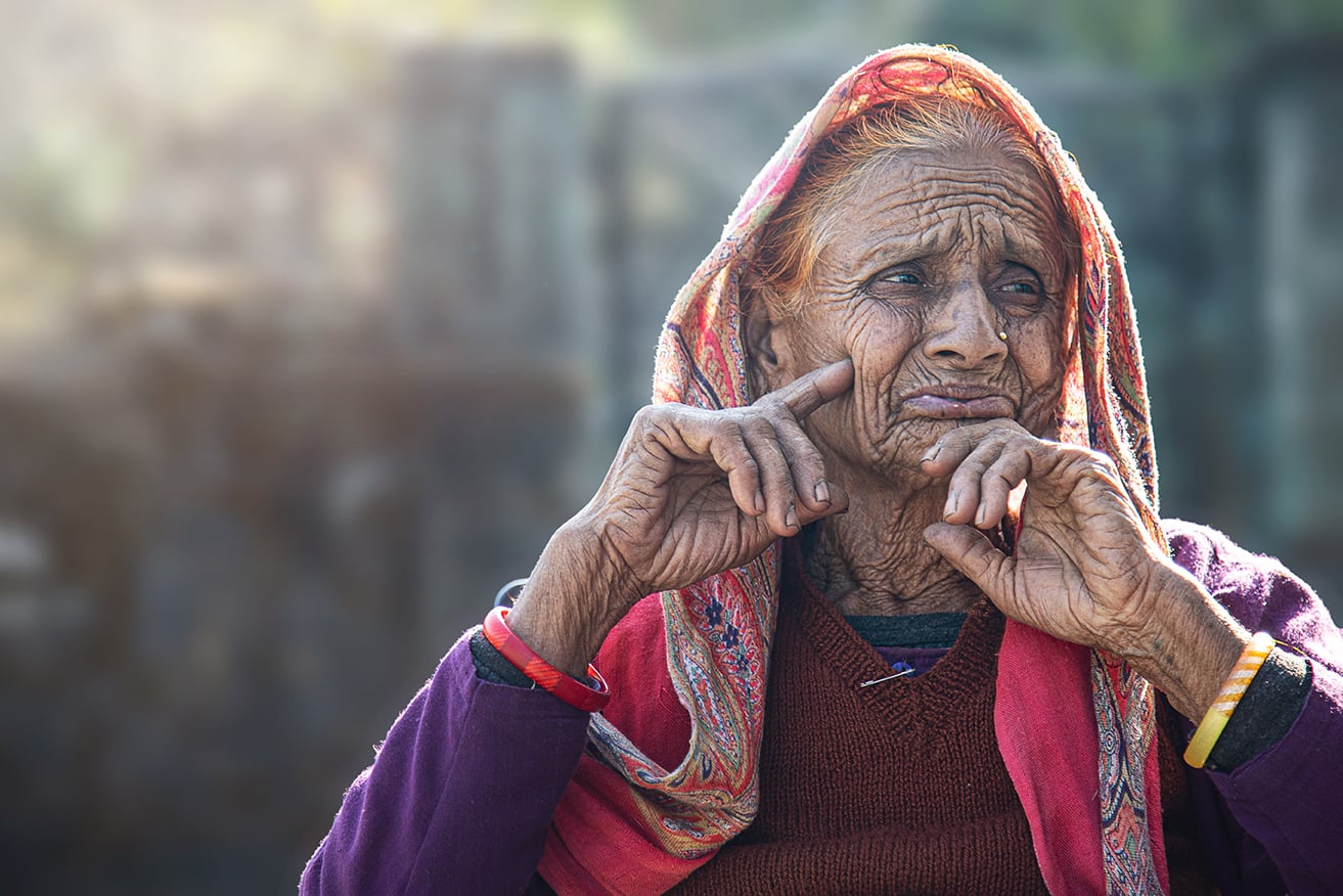 Elderly woman in a colorful scarf and sweater, resting her chin on her hand in a thoughtful pose, with deep wrinkles visible on her face.