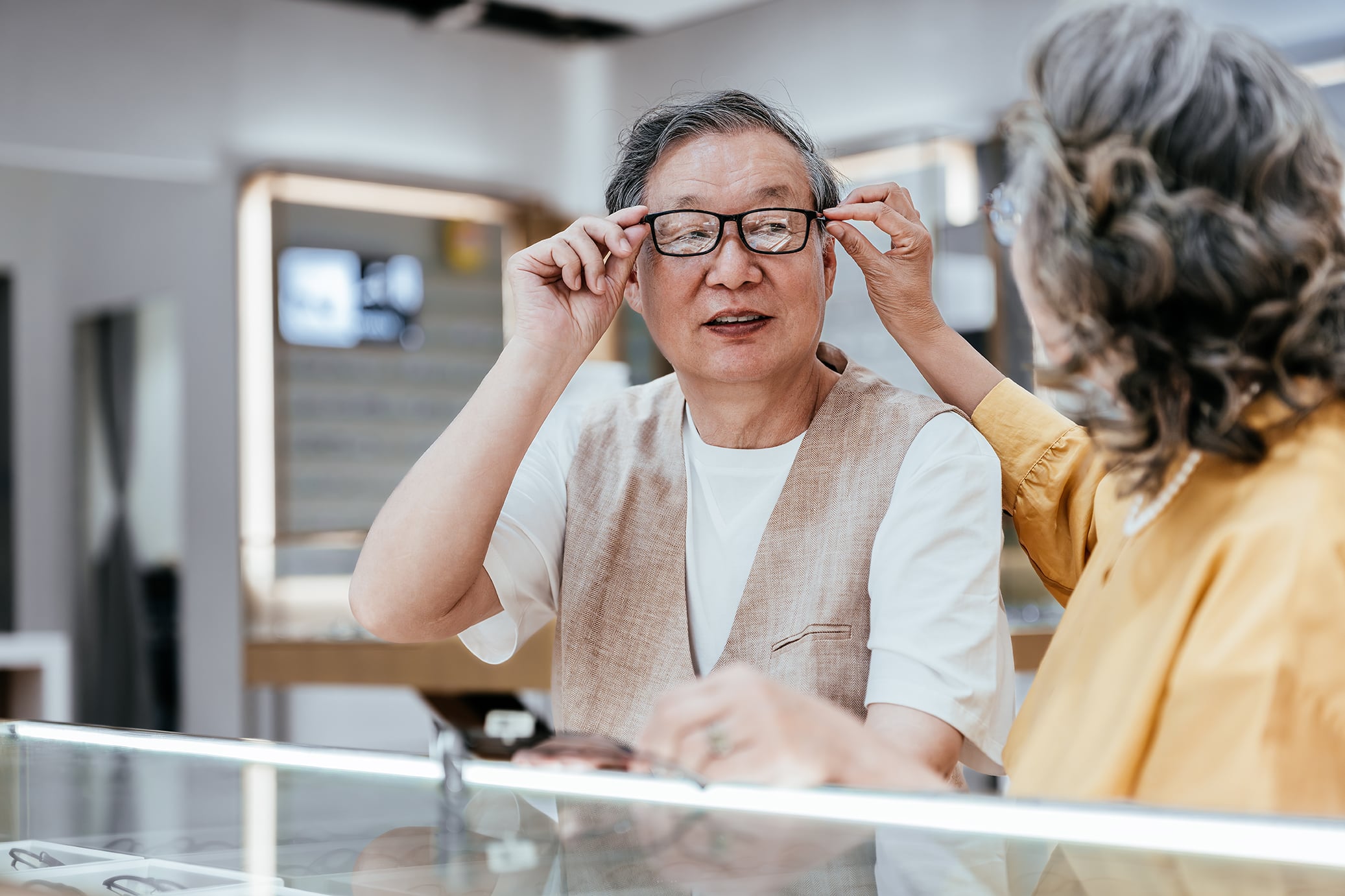 An older man trying on glasses in an optical store, assisted by a woman in a yellow blouse.