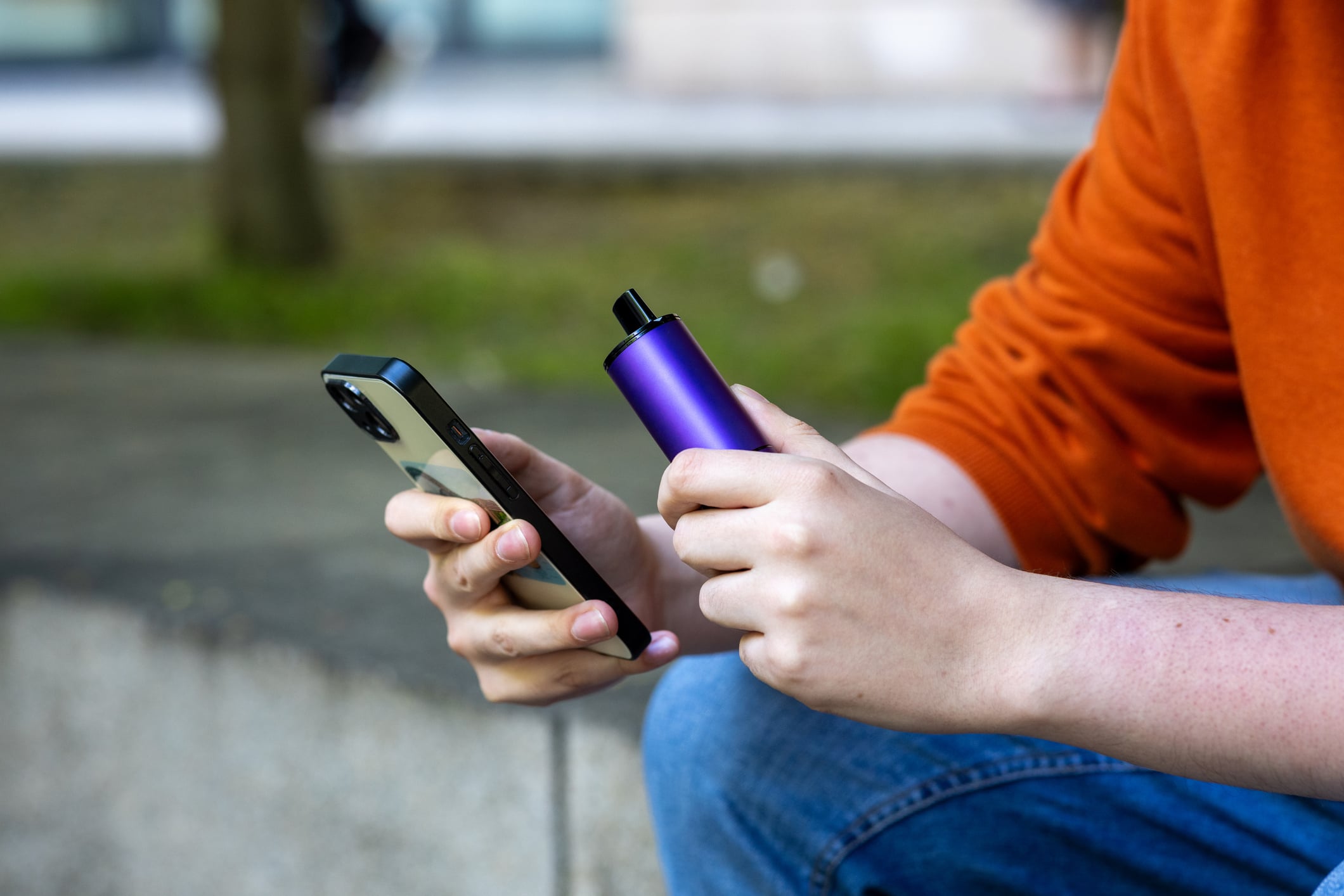 Young man vaping in Newcastle City Centre, sitting on a wall in a public area while he uses his mobile phone. He is using a disposable plastic vape pen.