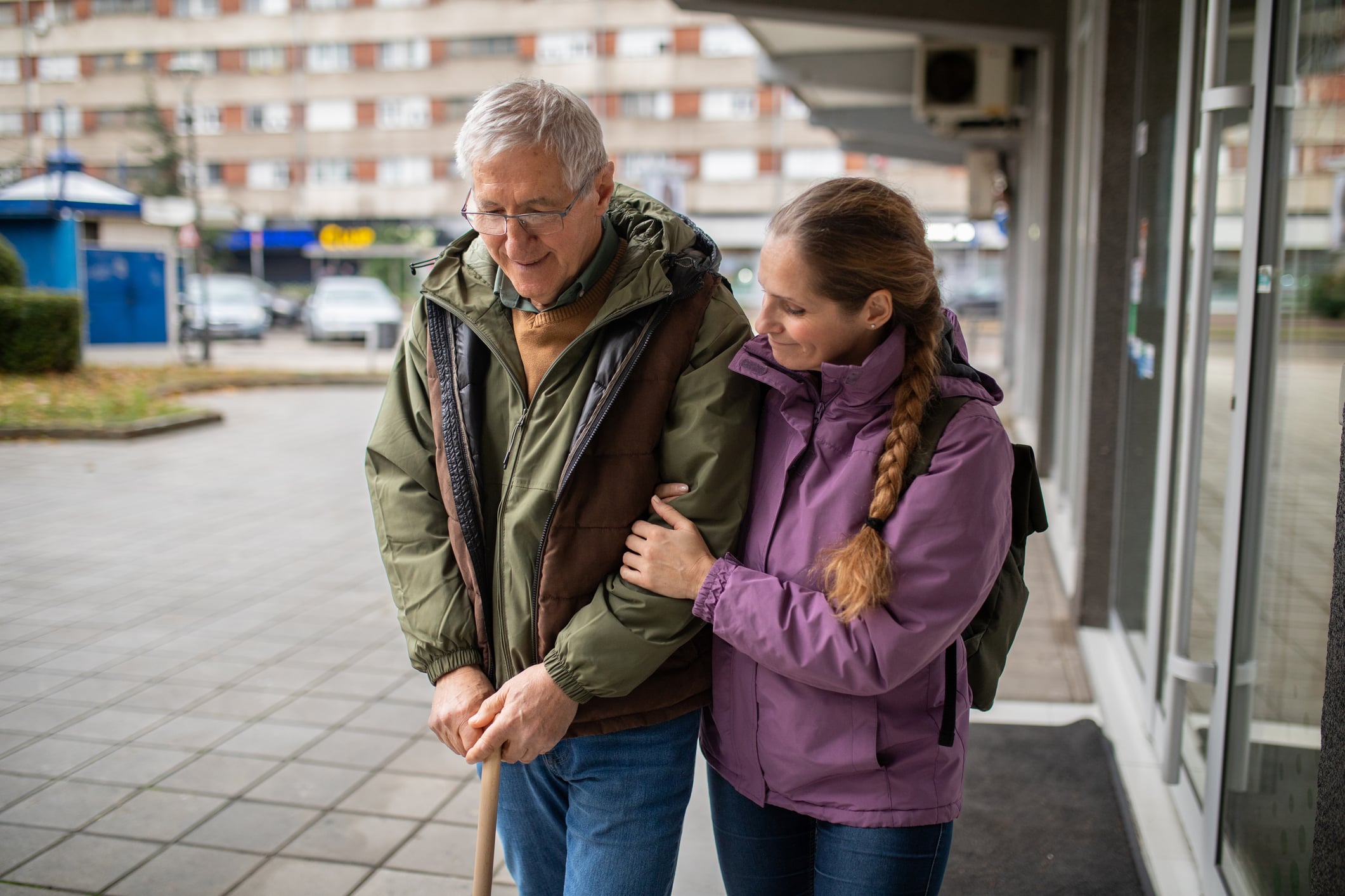 A mid adult Caucasian woman helping a senior man leave a hospital in a city